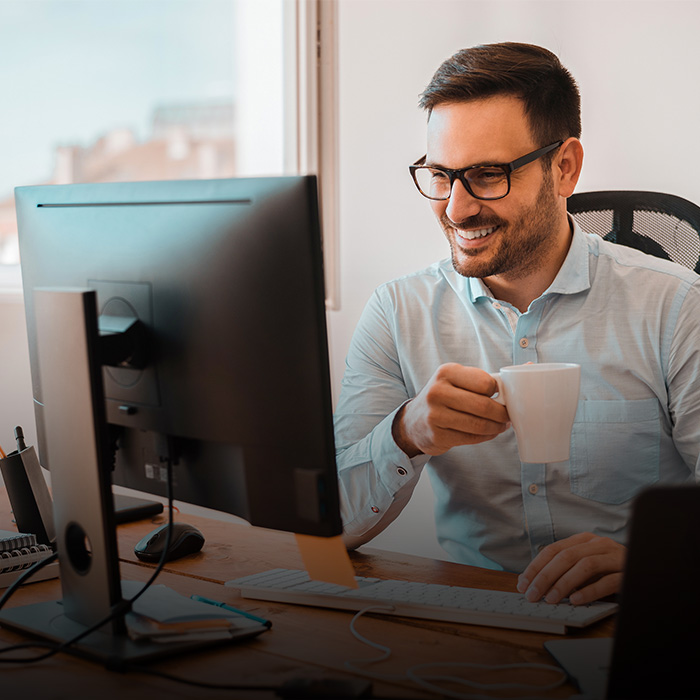 The picture shows a smiling man with a coffee cup in his hand working at the computer.